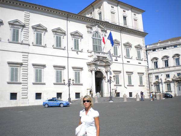 Blonde woman in white in front of the Quirinale Palace with flags