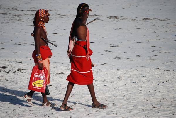 Two young Maasai men in red garments walk on white sand