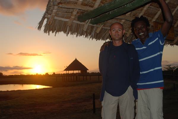 Two men hugging under a thatched hut as the sun rises