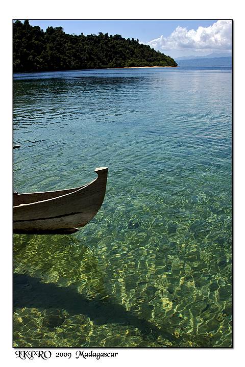 Wooden dugout canoe on clear green water, forested shore and beach, Madagascar