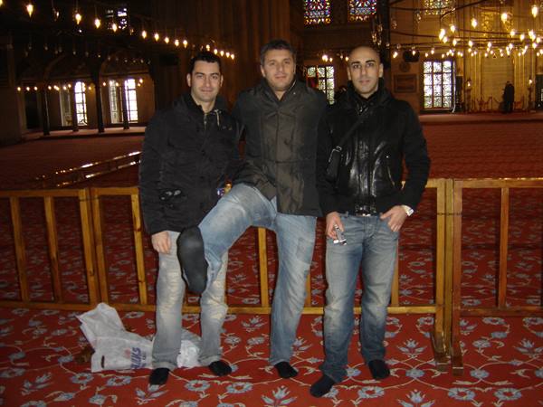 Three friends pose barefoot on a red carpet inside a mosque with hanging lamps
