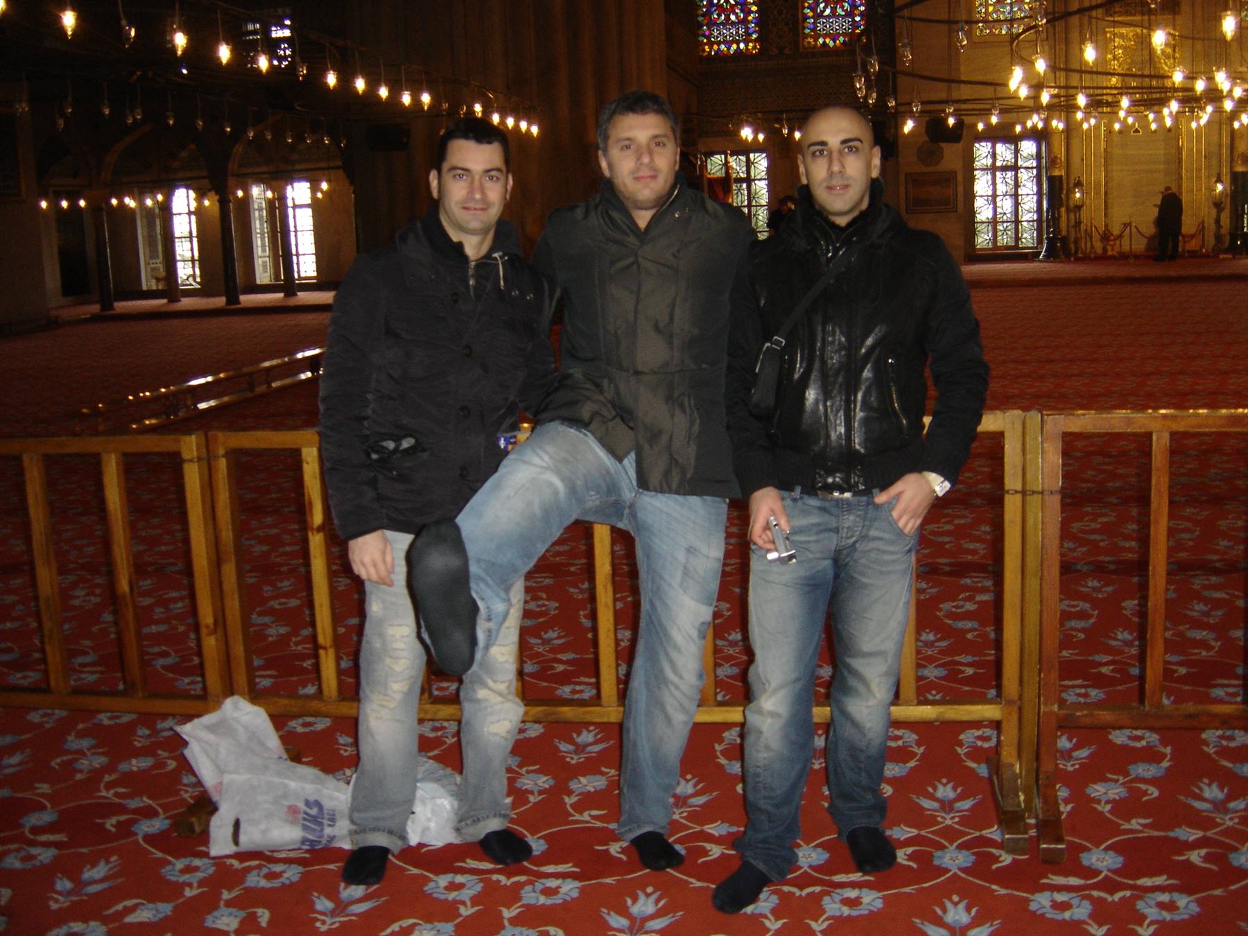 Three friends pose barefoot on a red carpet inside a mosque with hanging lamps