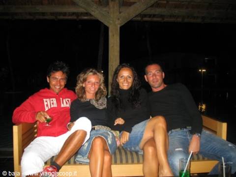 Four smiling friends on a wooden bench at night with drinks
