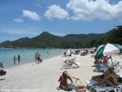 Beach with umbrellas, tourists on loungers, fruit vendor, green hills behind