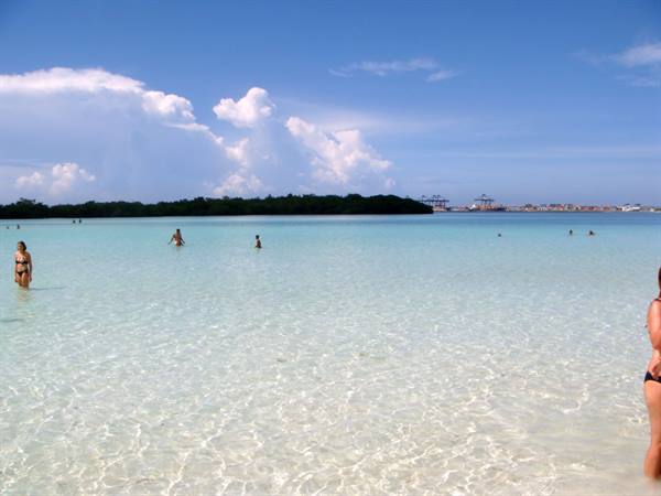 Swimmers in clear turquoise water, pale sand, container port in background