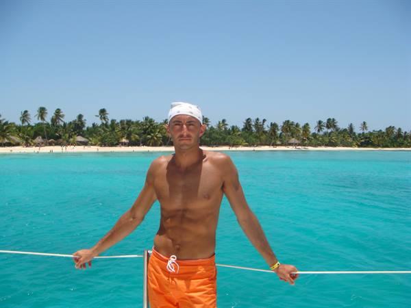 Man in orange swim shorts on a boat in front of a tropical palm beach