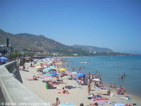 Crowded beach with colorful umbrellas, swimmers in turquoise sea, green hills behind