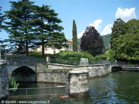 Stone walls along Como lakefront, people crossing the bridge among tall trees