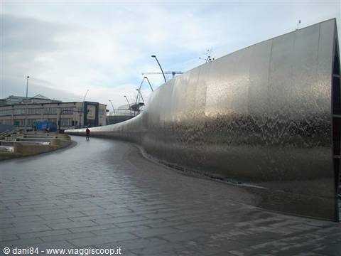 Large curved polished steel sculpture along a paved walkway under cloudy sky