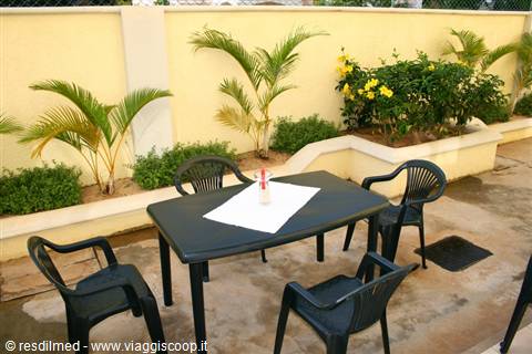 Black table with plastic chairs in courtyard, palm plants and yellow flowers along cream wall