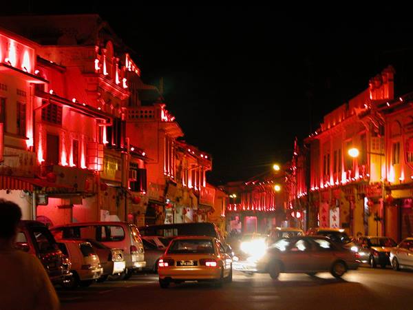 Cars pass at night between red-lit colonial shophouses, Jonker Street, Malacca
