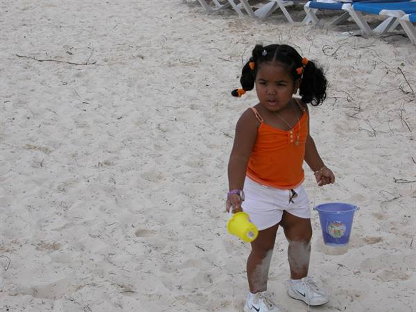 Little girl in orange top with blue bucket and yellow toy on the sand