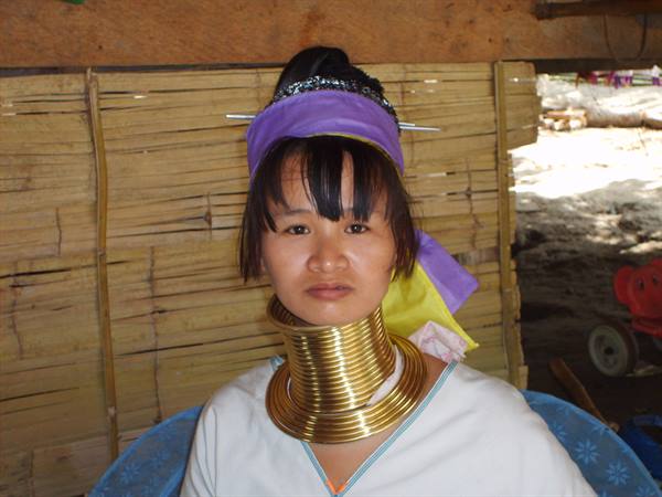 Kayan woman wearing brass neck coils and purple headscarf, bamboo wall behind