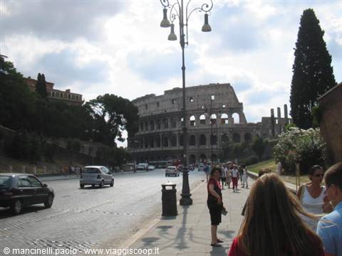 Tourists walk on the cobblestone street in front of the Colosseum under a cloudy sky