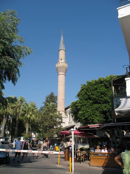 Stone minaret among palm trees and outdoor cafés, pedestrians in the sun