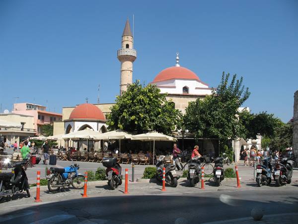 Mosque with minaret and outdoor cafés, parked scooters under sunny sky, Rhodes