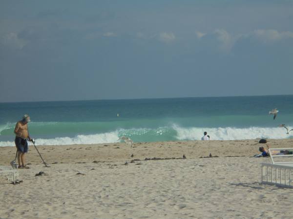 Man with metal detector on sand, turquoise waves and swimmers in the ocean