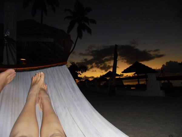 Feet on white hammock, dusk sky, palms and thatched roofs