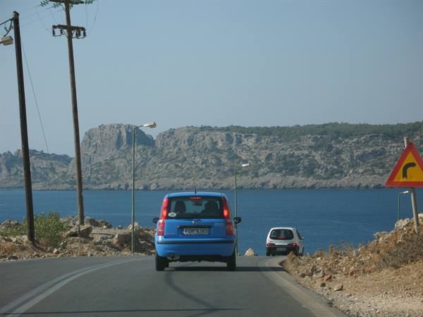 blue car on coastal road, rocky cliffs and blue sea, curve sign