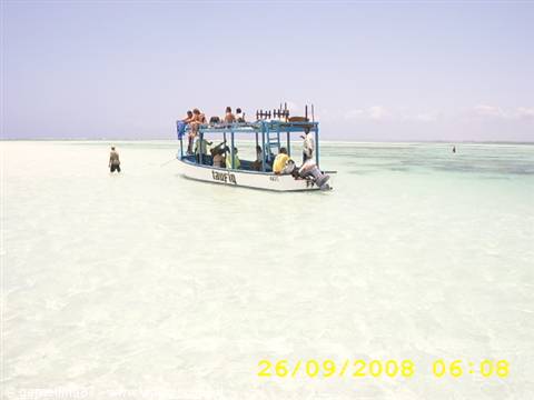 Blue boat with tourists anchored in shallow turquoise water, one person wading on white sand