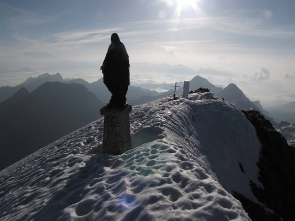 Madonna statue on snowy summit, high sun over mountain ridges