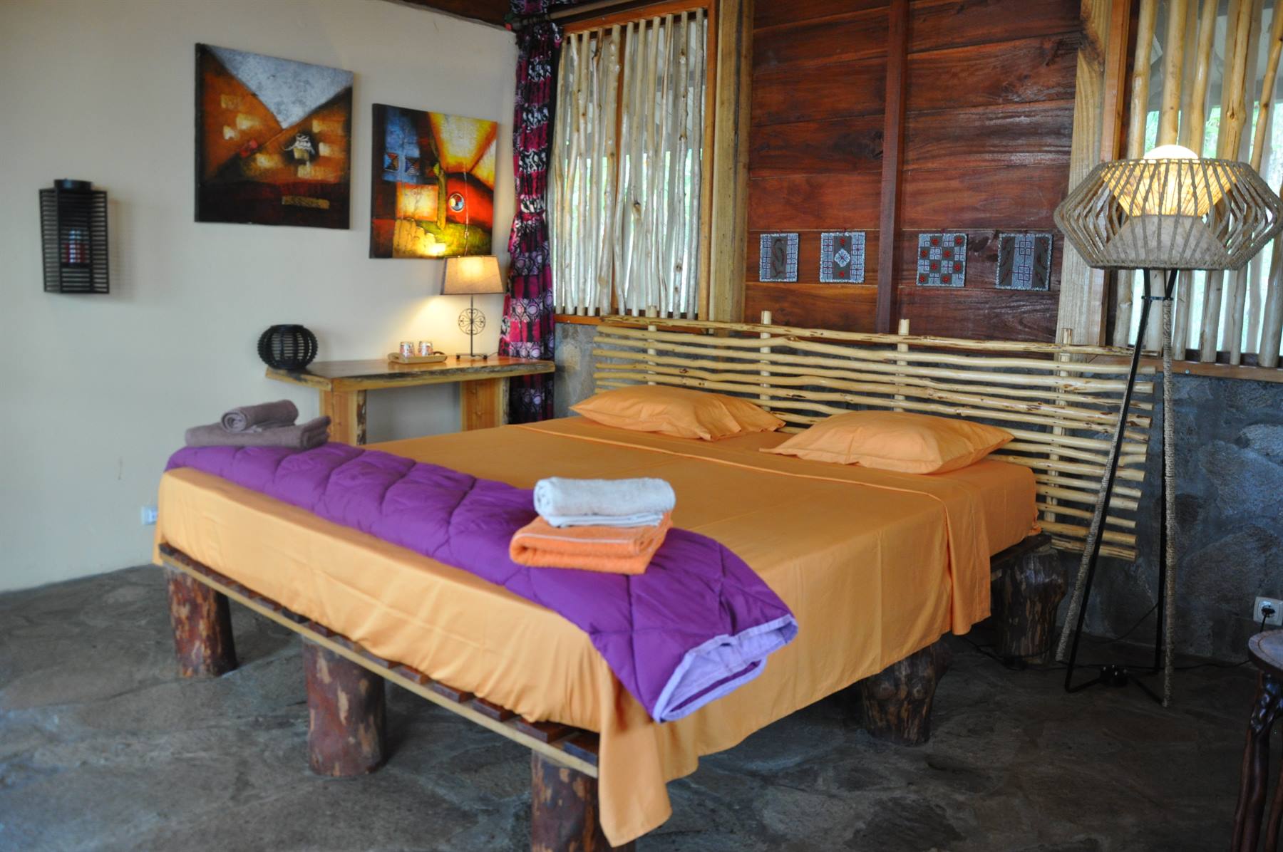 Camera con letto in legno su tronchi, lenzuola arancioni e coperta viola