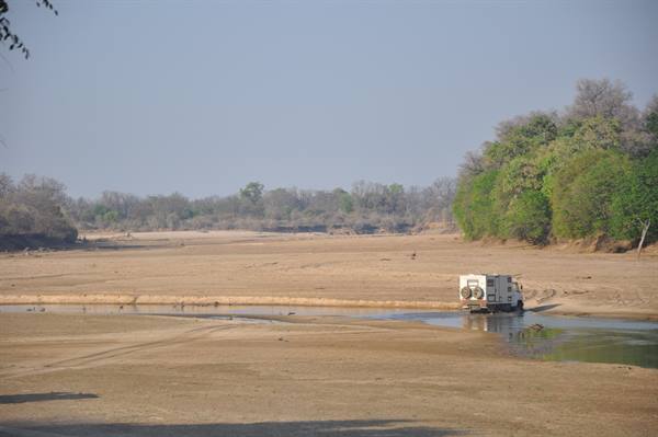 Overland truck crossing a dry sandy riverbed with shallow pools and green trees