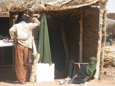 Man with green turban sitting under thatched hut, another at market stall