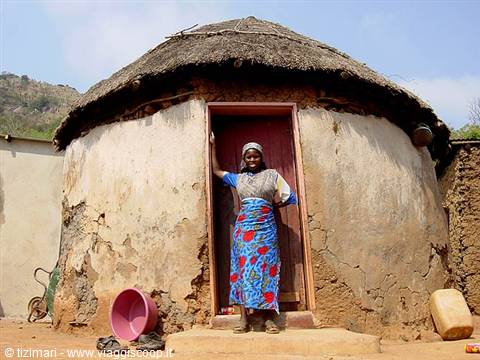 Woman in blue floral skirt smiles at the door of a round thatched mud hut