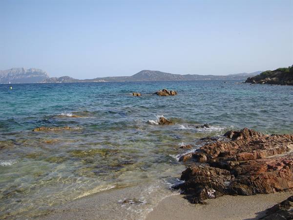 Red rocks on sandy beach, turquoise sea and distant islands under clear sky