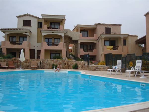 Blue pool in front of a resort with balconies, white loungers and a seated person