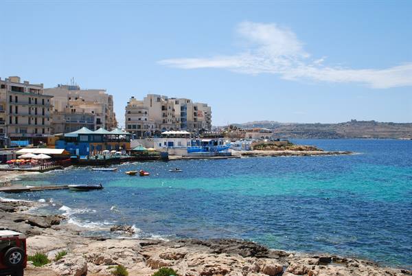 Buildings along rocky shore, boats and umbrellas on turquoise water, Malta