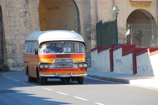 Vintage yellow and white bus drives past a stone gateway, Malta