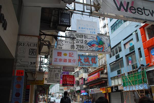 Layered neon signs above a busy Hong Kong street, pedestrians and bamboo scaffolding
