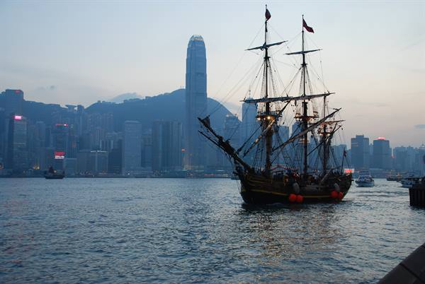 Three-masted tall ship in Hong Kong harbor at dusk, skyscrapers behind