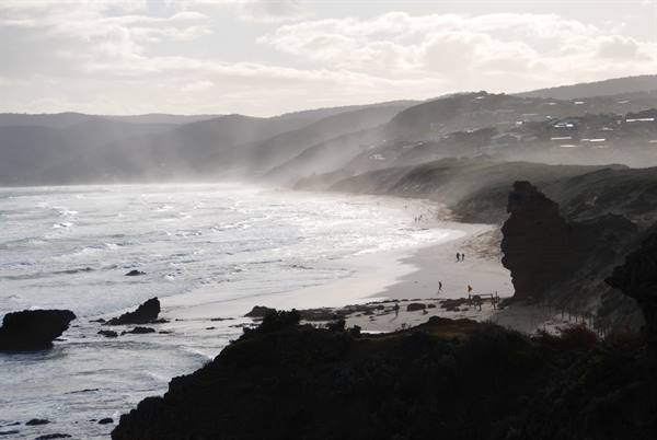 Spruzzi e nebbia marina sulla spiaggia di Apollo Bay, scogliere scure, case sulla collina