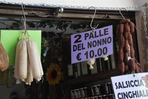 Cured meats hanging at a market stall, sign reads '2 palle del nonno €10'