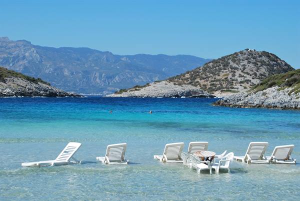 White sun loungers half-submerged in turquoise water, rocky hills and swimmers behind