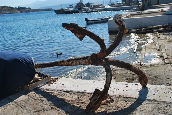Rusty four-armed anchor on the stone pier, boats and ferry in the harbor