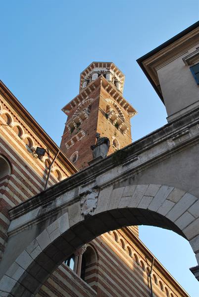Lamberti Tower in Verona seen through brick arch, warm sunlight, blue sky