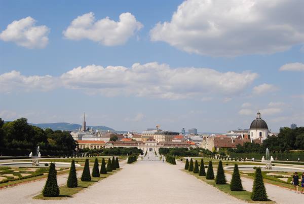Gravel path lined with conifers toward Belvedere Palace, cloudy sky over Vienna