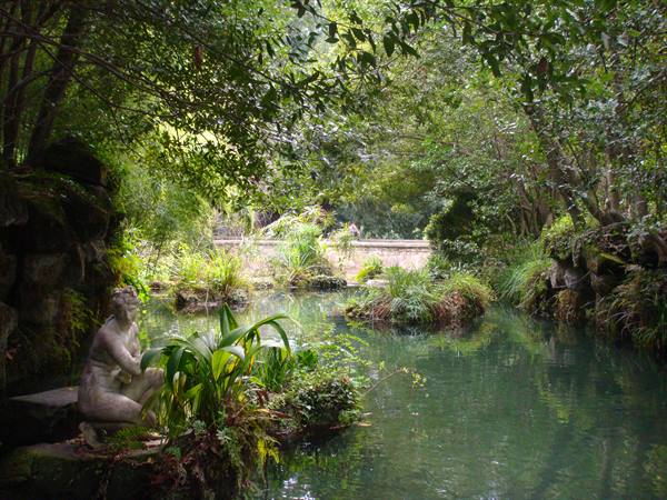 Stone statue beside a green pond surrounded by plants and trees in a garden
