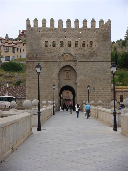 Tourists walk across the stone bridge toward a medieval fortified gate under gray sky