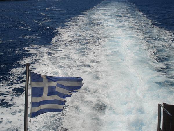 Greek flag waving over the foamy wake of a ferry on blue sea