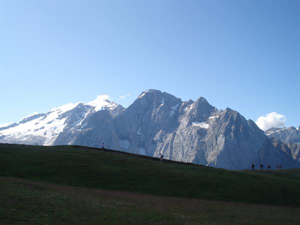 Hikers walk on green meadow in front of snow-dusted rocky peaks, blue sky