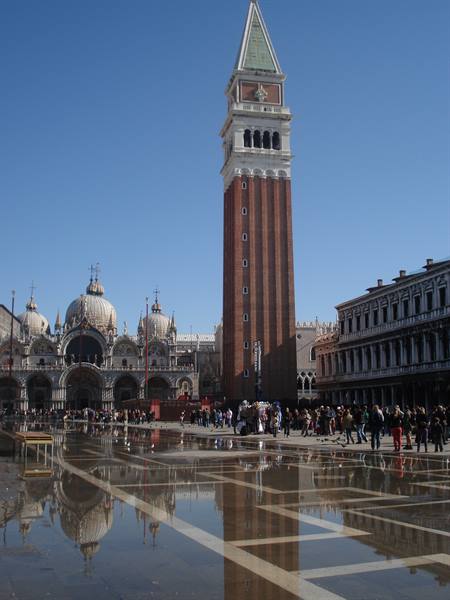 Bell tower and basilica in Venice, tourists standing on floodwater, bright sunlight