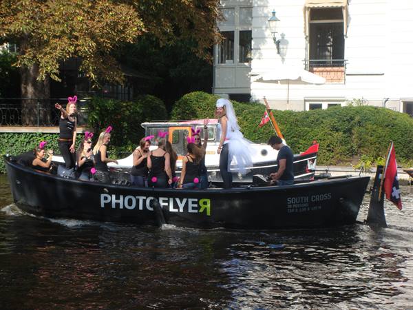 Group on black boat on Amsterdam canal, bride in veil waves