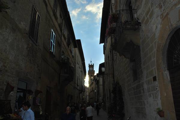 Stone medieval alley with tower in the distance, tourists walking among old houses
