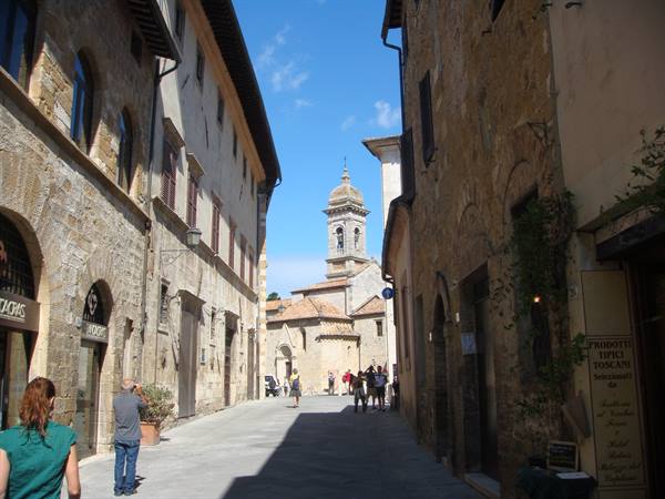 Tourists walk along the stone street of the village with baroque bell tower under sun