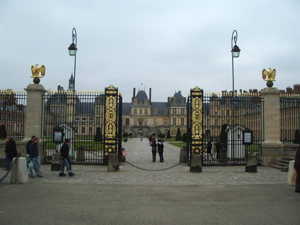 Gilded gates of the Château de Fontainebleau, tourists strolling under a grey sky